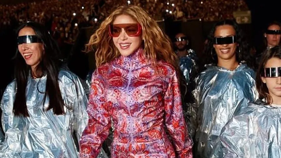 Shakira, con gafas de sol rojas y un mono estampado en rosa y morado, sonriendo mientras camina hacia el escenario junto a un grupo de 'Los Caminantes' con capas plateadas y gafas espejo, entre ellos artistas del Benidorm Fest