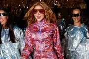 Shakira, con gafas de sol rojas y un mono estampado en rosa y morado, sonriendo mientras camina hacia el escenario junto a un grupo de 'Los Caminantes' con capas plateadas y gafas espejo, entre ellos artistas del Benidorm Fest