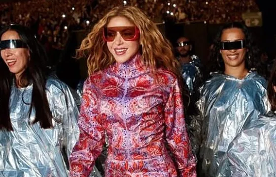 Shakira, con gafas de sol rojas y un mono estampado en rosa y morado, sonriendo mientras camina hacia el escenario junto a un grupo de 'Los Caminantes' con capas plateadas y gafas espejo, entre ellos artistas del Benidorm Fest